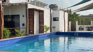 Una piscina con borde de azulejos, rodeada por una terraza de madera y un edificio blanco con antena parabólica.