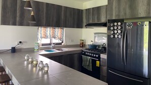 Una cocina moderna con un refrigerador de acero inoxidable, una estufa a gas y un lavamanos. Hay frascos en la encimera y una ventana con cortinas.