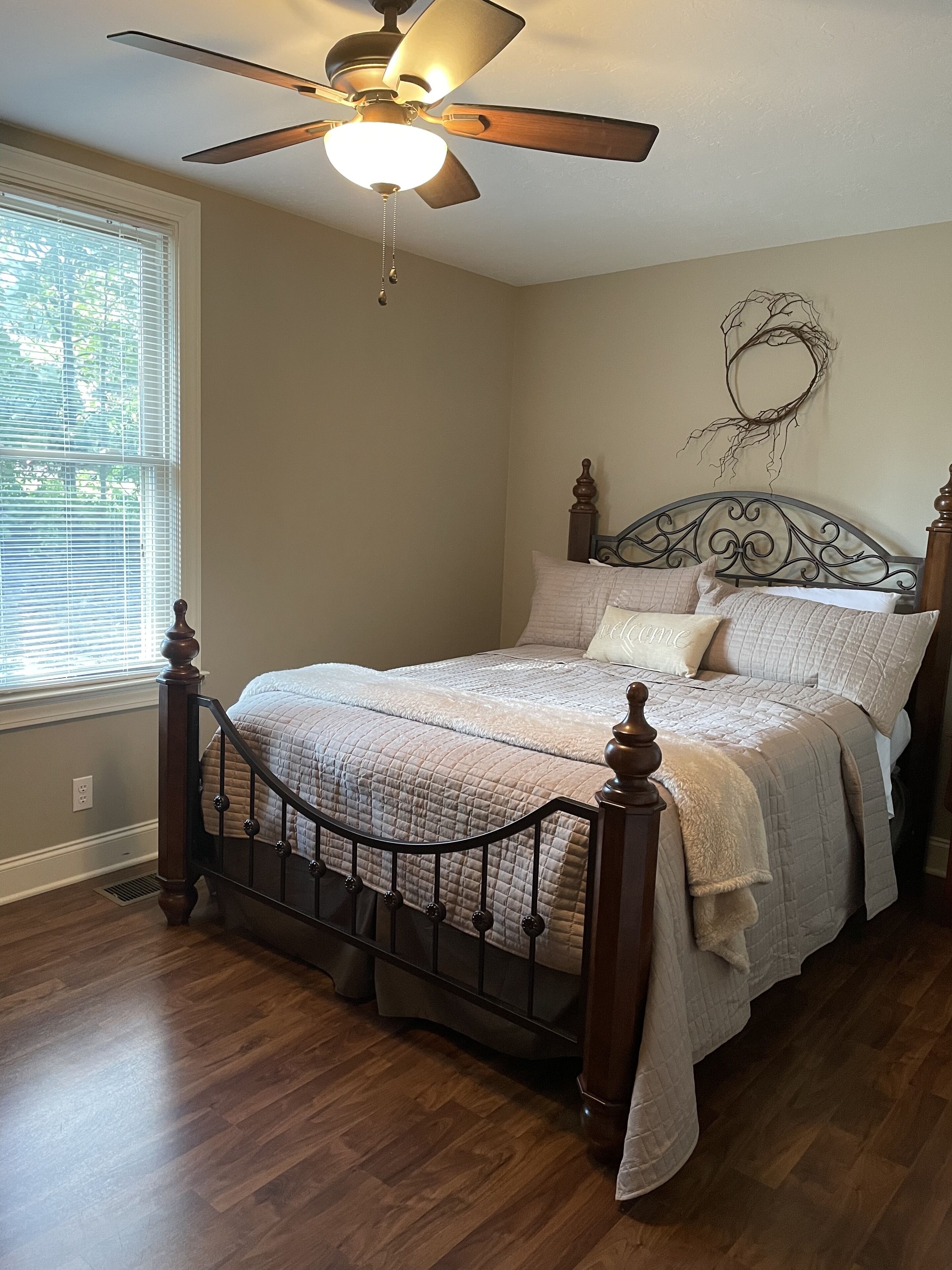 Quarto com cama, piso de madeira, ventilador de teto e janela com persianas.
