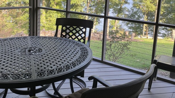 Un patio cubierto con una mesa redonda de metal y dos sillas de mimbre, ventanas amplias con vistas a un paisaje verde.