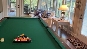 Una mesa de billar con bolas, un ventilador de techo y vistas al exterior a través de amplios ventanales.