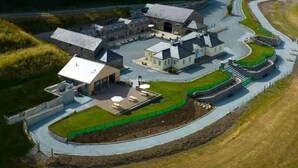 An aerial view of a large, historic building complex with multiple structures, including a central building with a large roof and several smaller buildings. There is a paved driveway leading to the entrance, and the area is surrounded by grassy fields. - Magical 3-bed Stone Built Cottage - Sleeps 6 (Ballynahinch)