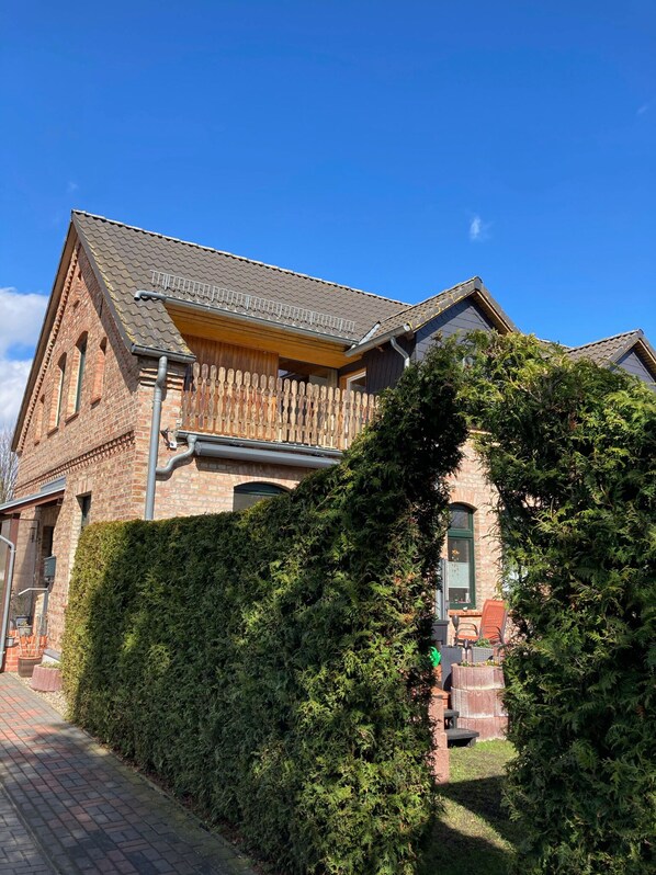 A brick house with a wooden balcony, a paved pathway, and a well-maintained hedge. - Vacation apartment on the Szczecin Lagoon with garden and terrace (Ueckermünde)