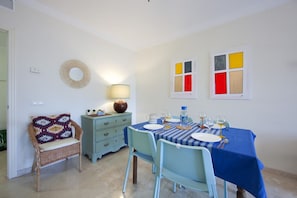 A dining area with a table set for four, a wicker chair, a blue tablecloth, and a wall with colorful artwork.