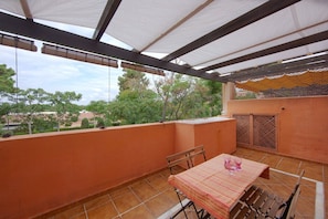 A covered outdoor patio with a table and chairs, a view of trees and buildings, and a pergola structure.