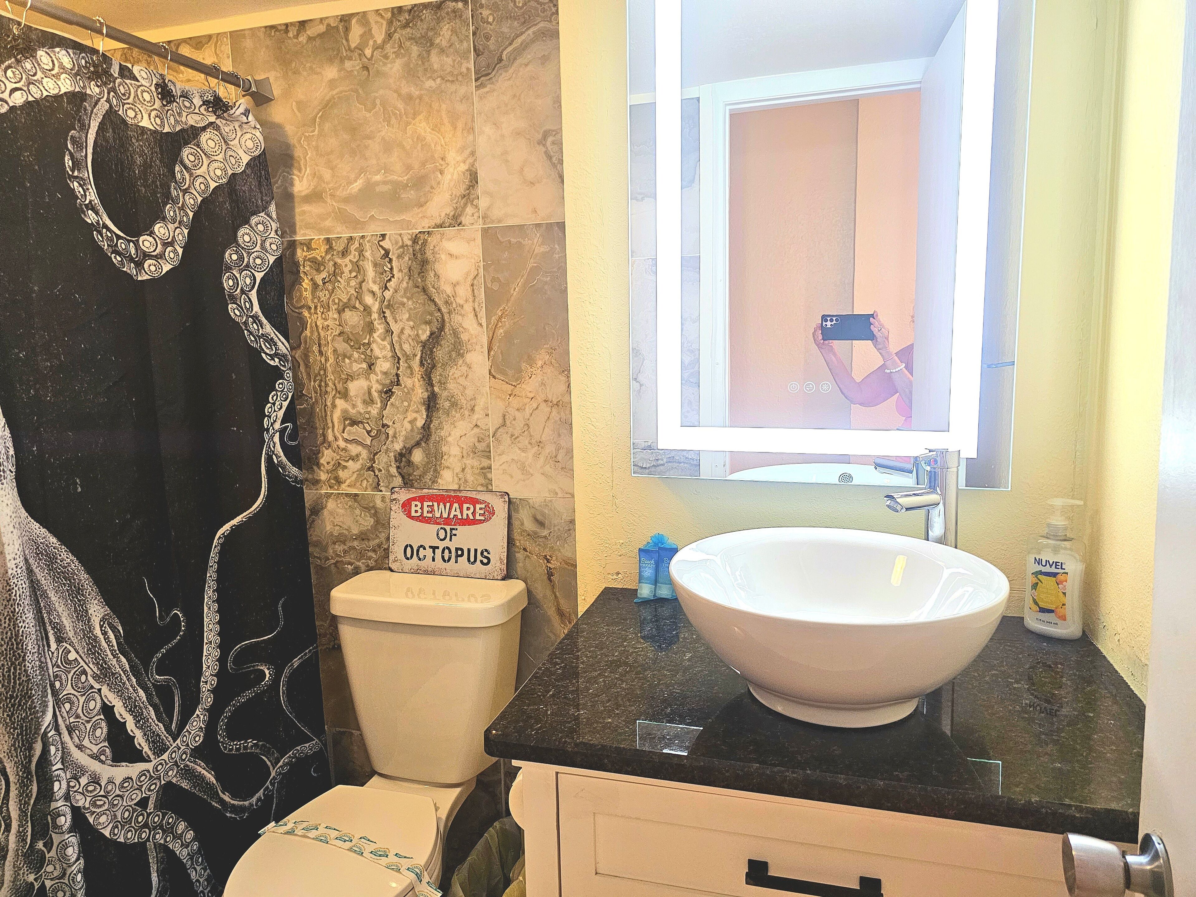Et badeværelse med et bruseforhæng med et sort-hvidt blæksprutte-tema, et toilet, en vask med en Nuvel håndspritflaske og et spejl, der afbilder en person, der tager et billede.
