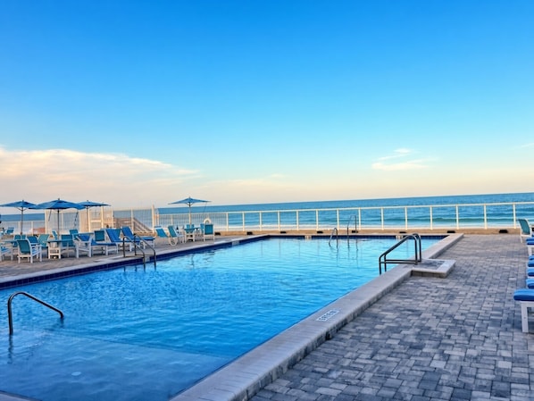 Una piscina con sillas de descanso y sombrillas, una barandilla blanca y vista al océano.
