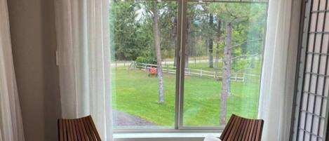 Een kamer met twee houten stoelen met witte kussens, een kleine ronde tafel en een groot raam met uitzicht op een grasveld en bomen.