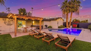 Un patio trasero con piscina, mobiliario exterior e iluminación colgante.