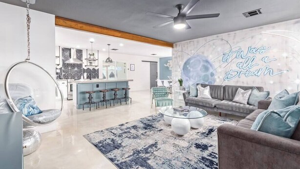 Una sala moderna con sofá, mesa de centro y una silla colgante. Se observa una zona de cocina al fondo con taburetes de bar.