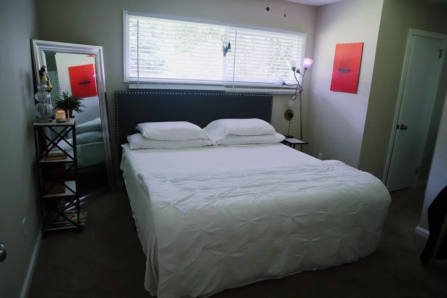 A bedroom with a large bed, a bedside table, a mirror, a window with blinds, and a door.