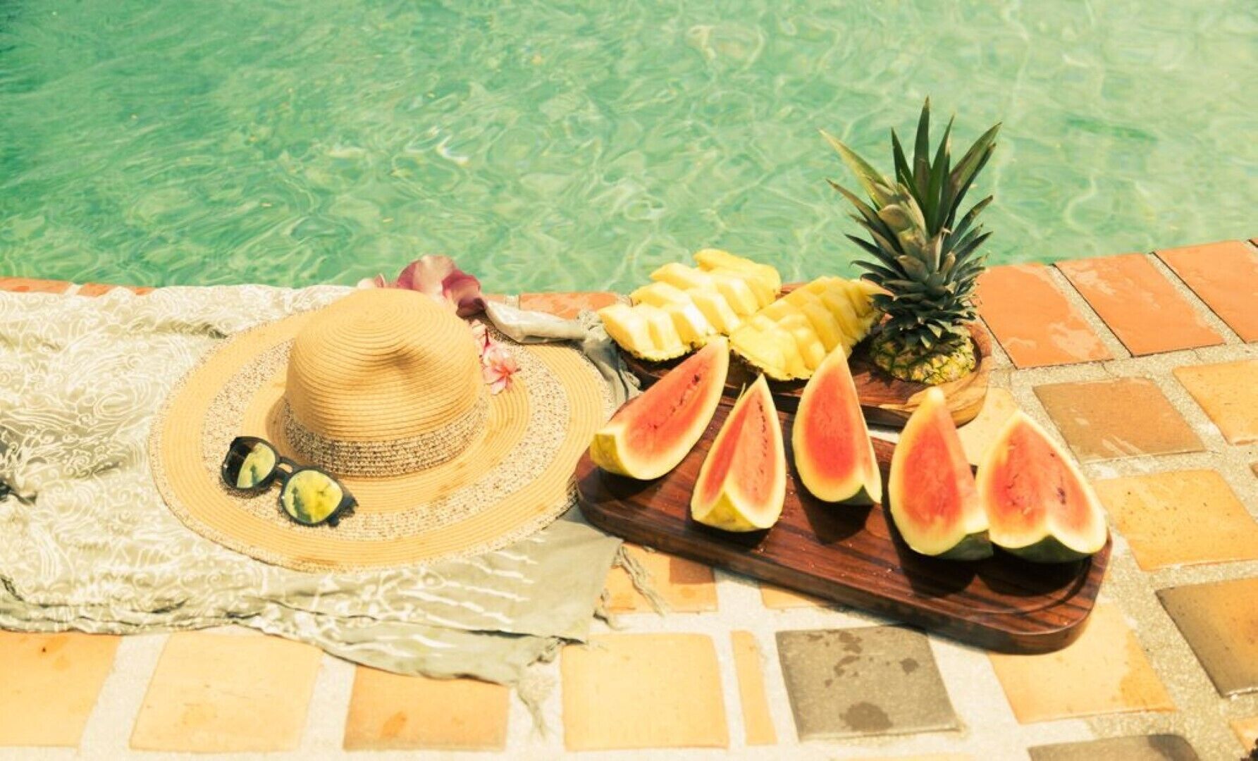 Une scène de bord de piscine avec un chapeau, des tranches de pastèque, de l’ananas et un plateau en bois.
