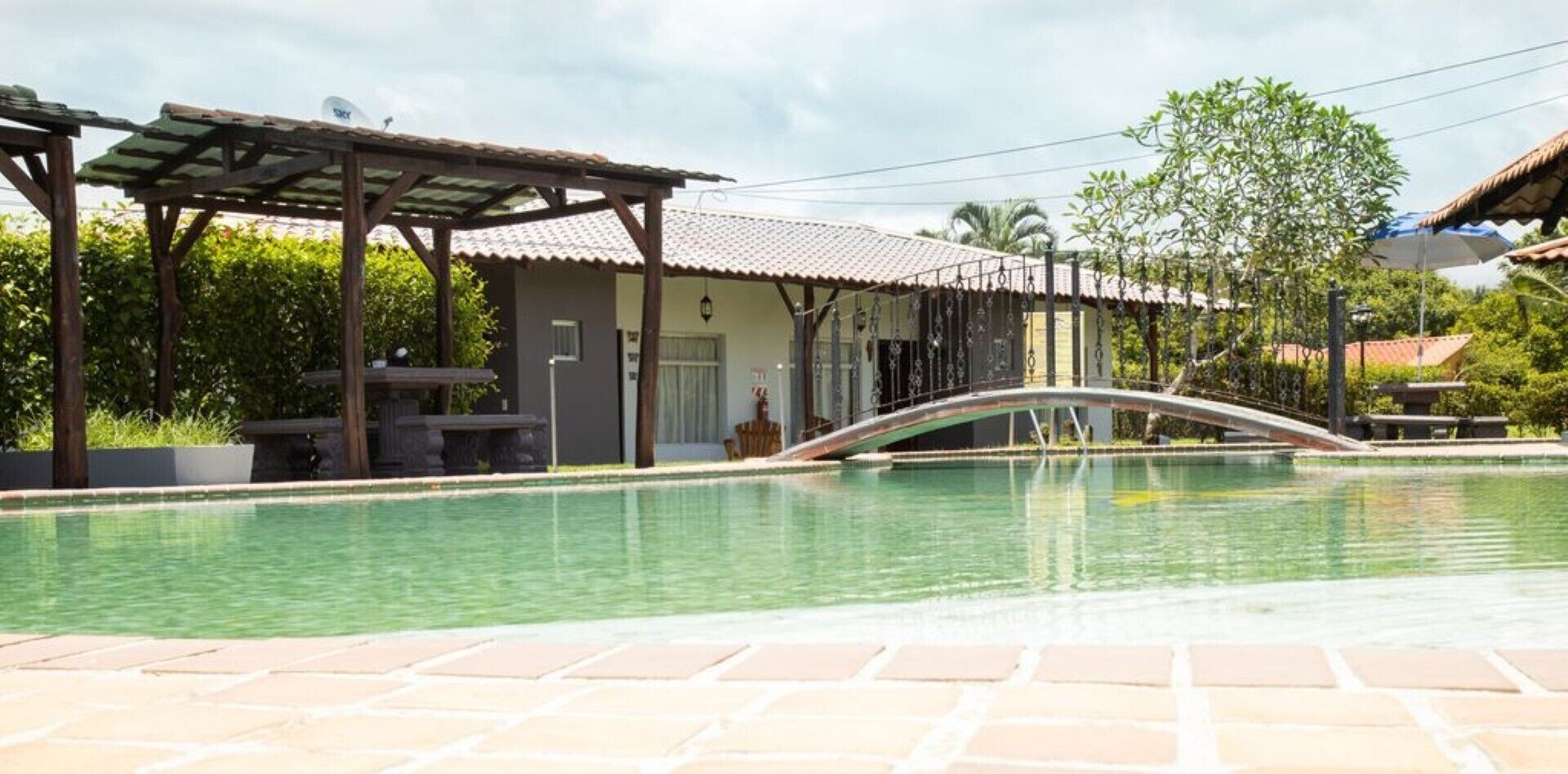 A swimming pool with a bridge, a covered patio, and a building with a tiled roof.