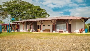 A two-story building with a covered patio area, a playground, and a large tree in the background. - Hotel Rancho Leon a Place to Stay | Room 12 (Quepos)