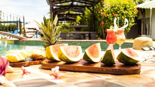 A tropical setting with a pool, fruit slices, and drinks on a table. - Hotel Rancho Leon a Place to Stay | Room 12 (Quepos)