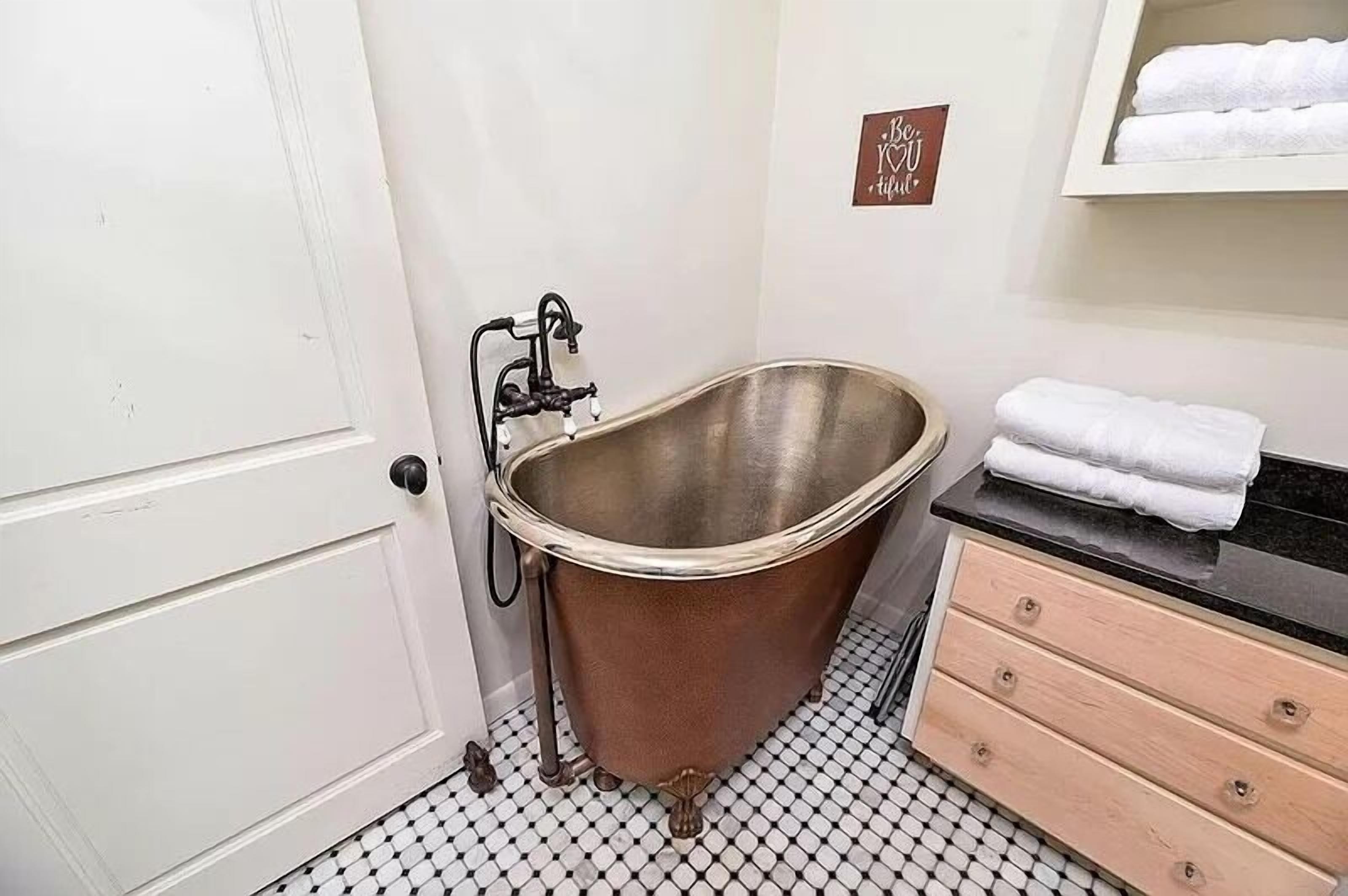 A bathroom with a vintage clawfoot bathtub, a white door, a black countertop with a towel rack, and a sign that says "Be Kind."