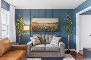 Un salon avec un canapé gris, deux coussins à motifs, un tableau représentant un champ et deux grandes plantes.