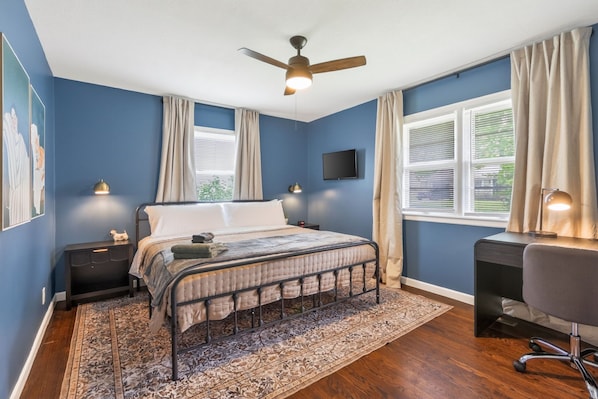 A bedroom with a bed, a desk, a chair, a ceiling fan, a TV, and a window with curtains.