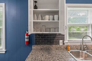 A kitchen with a blue wall, a white cabinet with glassware, a granite countertop, and a stainless steel sink.