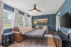 A bedroom with a bed, two sofas, a TV, a ceiling fan, and a window with curtains.