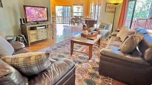 A spacious living room with a large leather sofa, a coffee table, and a television. There is a dining area with a table and chairs visible in the background.