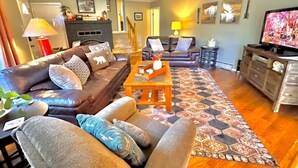 A living room with a sofa set, a coffee table, and a patterned rug. There is a fireplace, a television, and a staircase leading to another room.
