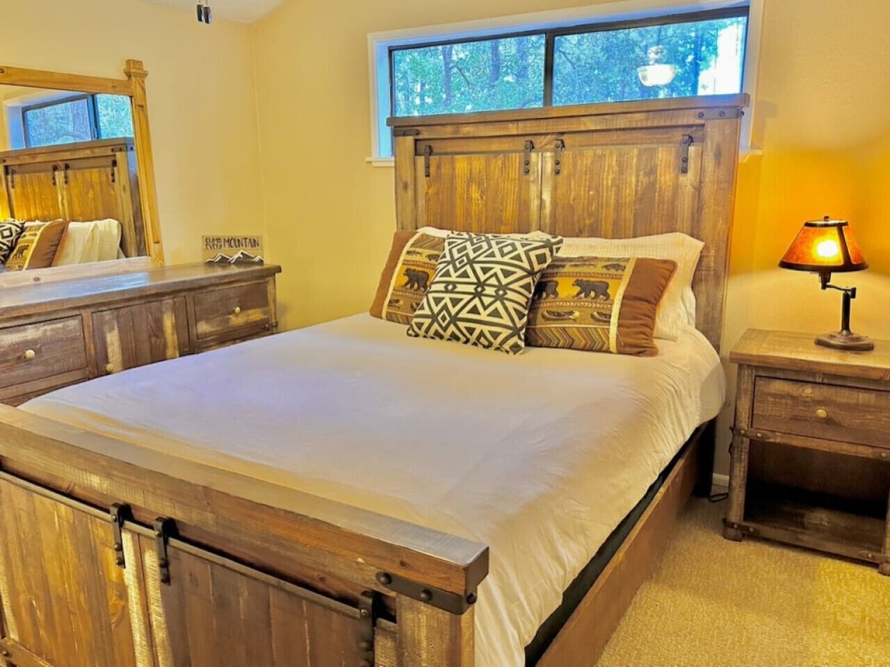 A wooden bed with a headboard, a nightstand with a lamp, and a window with a view of trees.