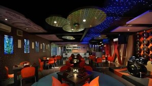 Un restaurante con una mesa redonda central rodeada de sillas rojas, una zona de bar al fondo y luces decorativas en el techo.