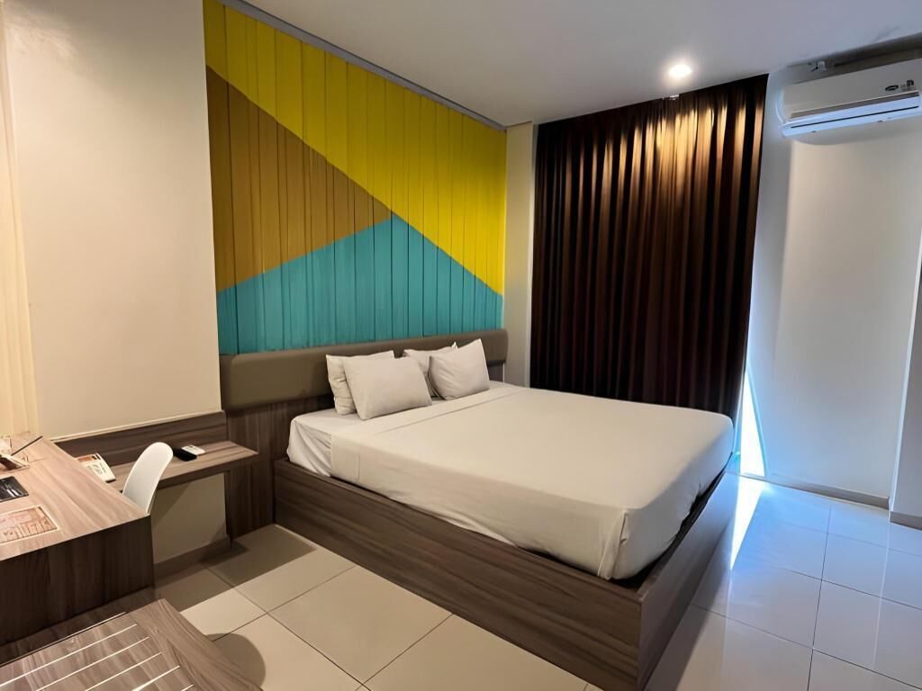 Ein Hotelzimmer mit einem Bett, einem Schreibtisch, einem Stuhl, einer Wand mit geometrischem Muster, einem Fenster mit Vorhängen und einer Klimaanlage.