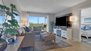 Un salon moderne avec un canapé, une table basse et un téléviseur. Il y a un balcon avec vue sur l’extérieur.