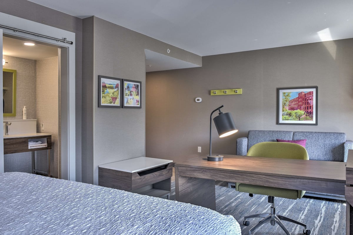 A modern hotel room with a bed, a desk, a chair, a lamp, and two framed pictures on the wall.