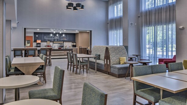 A modern dining area with a central seating area, a kitchen in the background, and large windows providing natural light. - Hampton Inn & Suites Keene (Keene)
