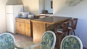 Una cocina con armarios de madera, un refrigerador, un microondas y un comedor con mesa de vidrio y sillas de mimbre.