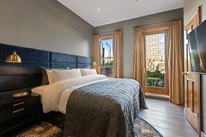Une chambre à coucher avec un grand lit, une table de chevet, une lampe et une vue sur un immeuble par la fenêtre.