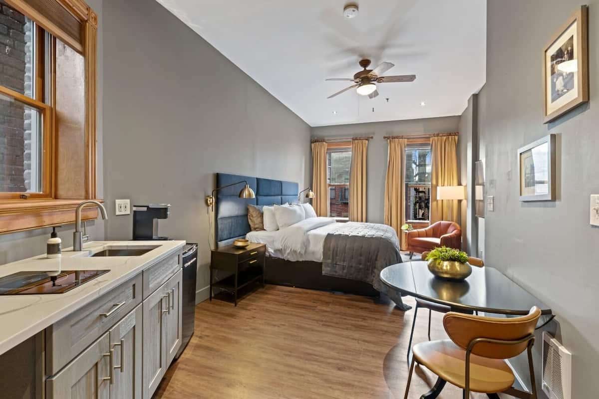 Een kleine hotelkamer met kitchenette, een bed met blauw hoofdeinde, een eettafel met stoelen en een plafondventilator.