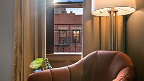 Une pièce d’angle avec un fauteuil marron, un lampadaire et une fenêtre donnant sur un immeuble en briques.