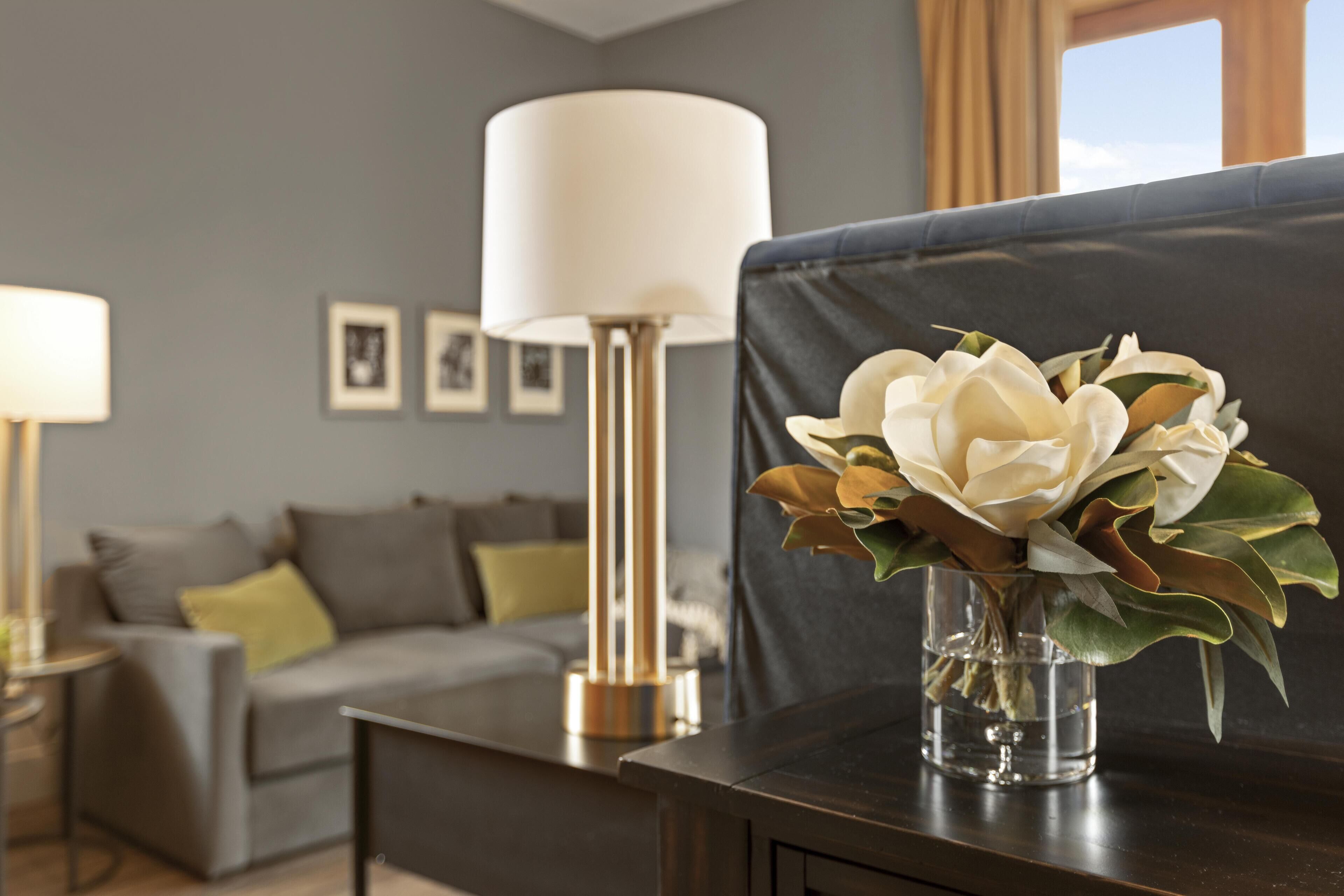 Una sala de estar moderna con un sofá gris, un jarrón de cristal con flores sobre una mesa de madera oscura y una lámpara de pie.
