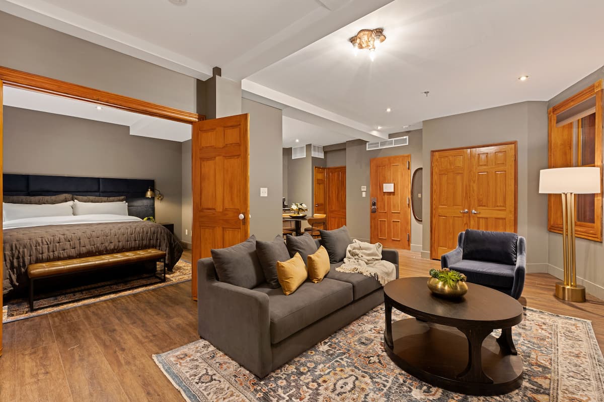 Een moderne hotelkamer met een groot bed, een bank, een salontafel en een stoel. Er zijn houten deuren en een kroonluchter.