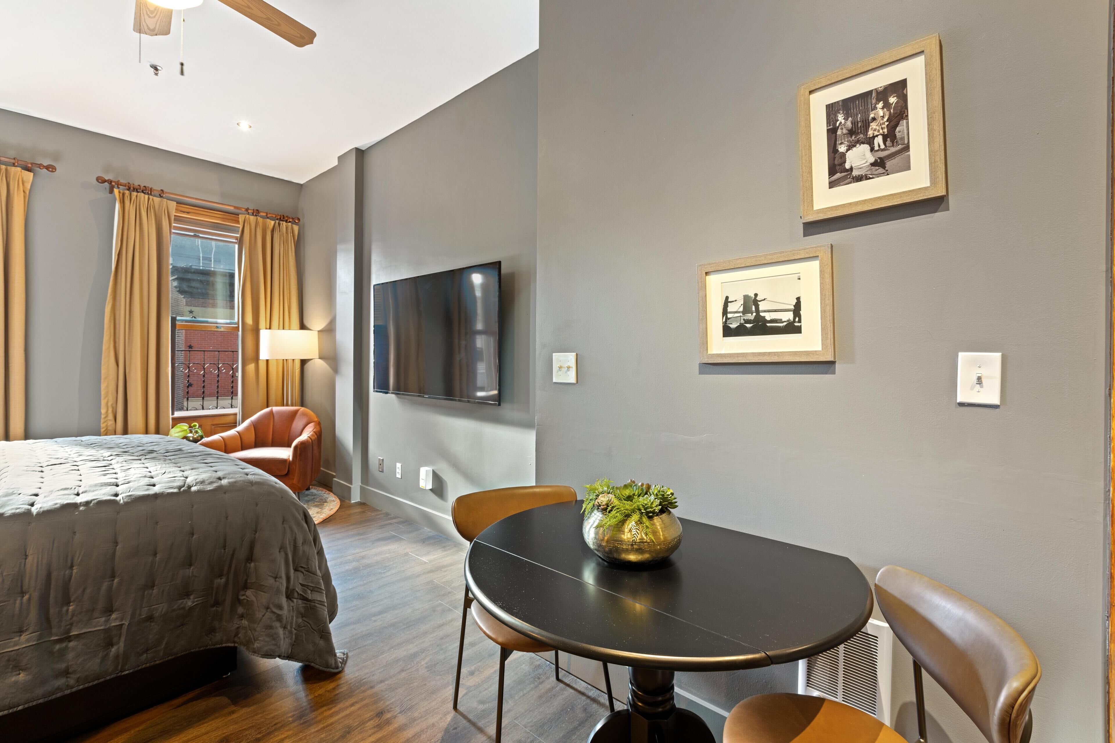 A modern hotel room with a bed, a dining table with chairs, a television, and framed pictures on the wall.