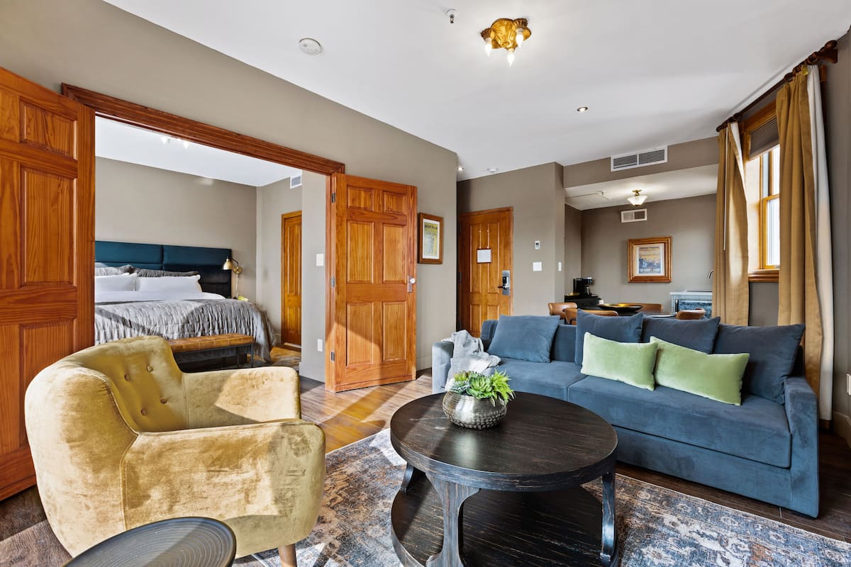Een hotelkamer met een bed, een blauwe bank, een gele fauteuil, een ronde salontafel en houten deuren.