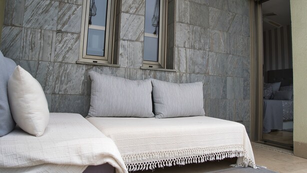 Un letto con copriletto bianco e cuscini grigi, appoggiato a un muro in pietra con due finestre.