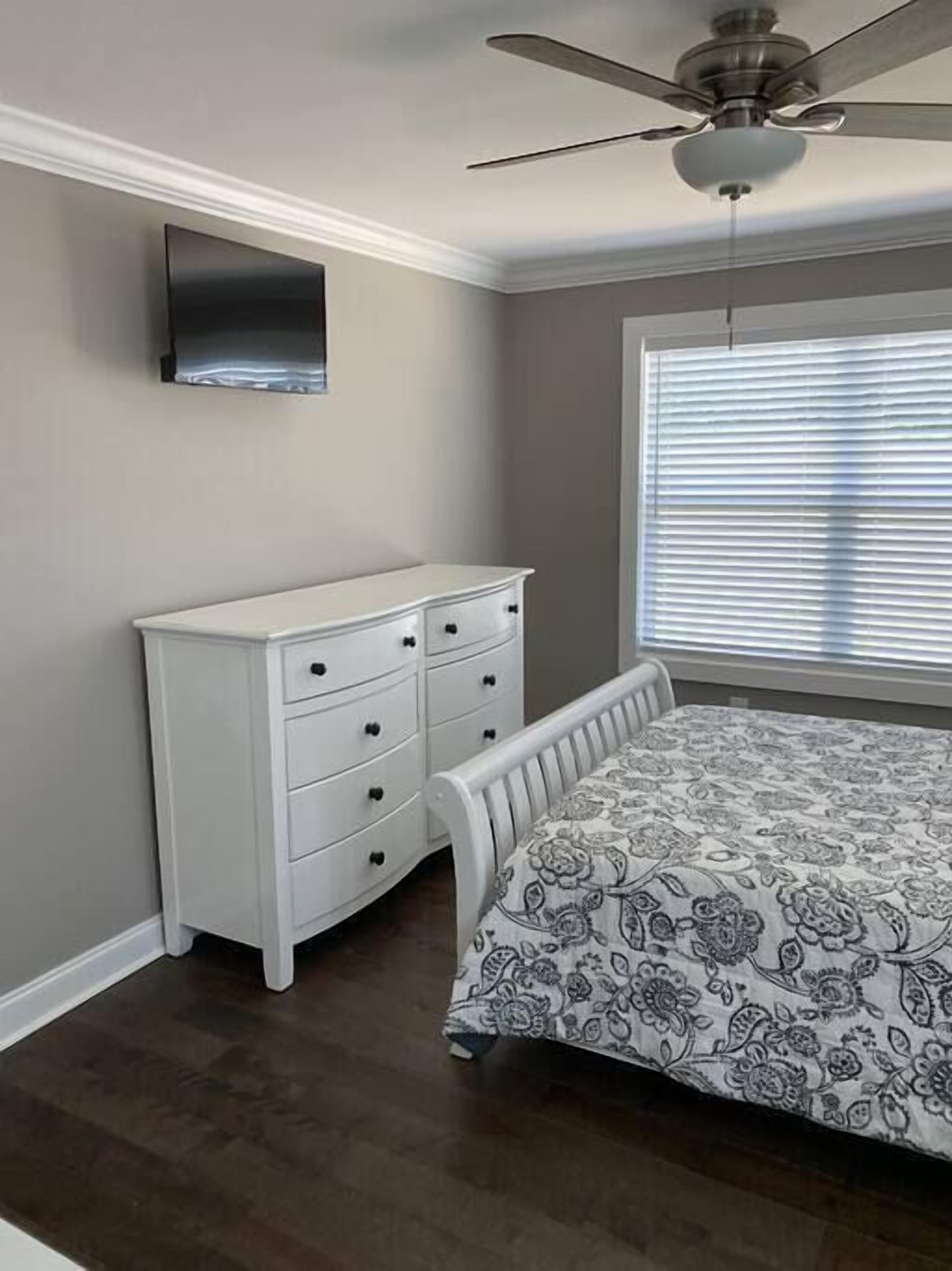 Un dormitorio con cama, cómoda, ventilador de techo y ventana con persianas.