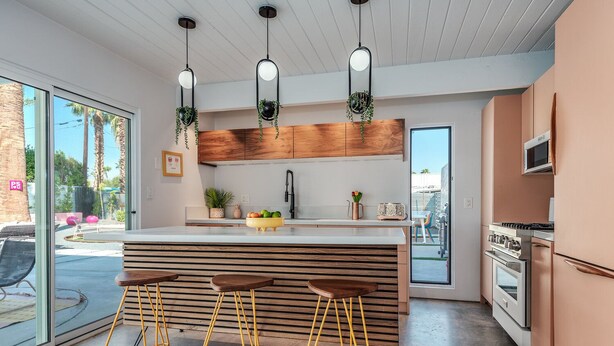 A modern kitchen with a central island, bar stools, hanging plants, and a view of the outdoors. - Dreamy Mid Century Modern House with Saltwater Pool &  Resort Style Backyard (Palm Springs)