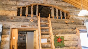 Interior de una cabaña de troncos con una escalera de madera que lleva a un altillo, una puerta con un pequeño ventanal y una decoración festiva en la pared.
