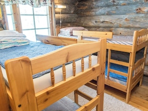 Una cama litera de madera con cabecera y piecera, una mesita de madera y una ventana con cortinas.