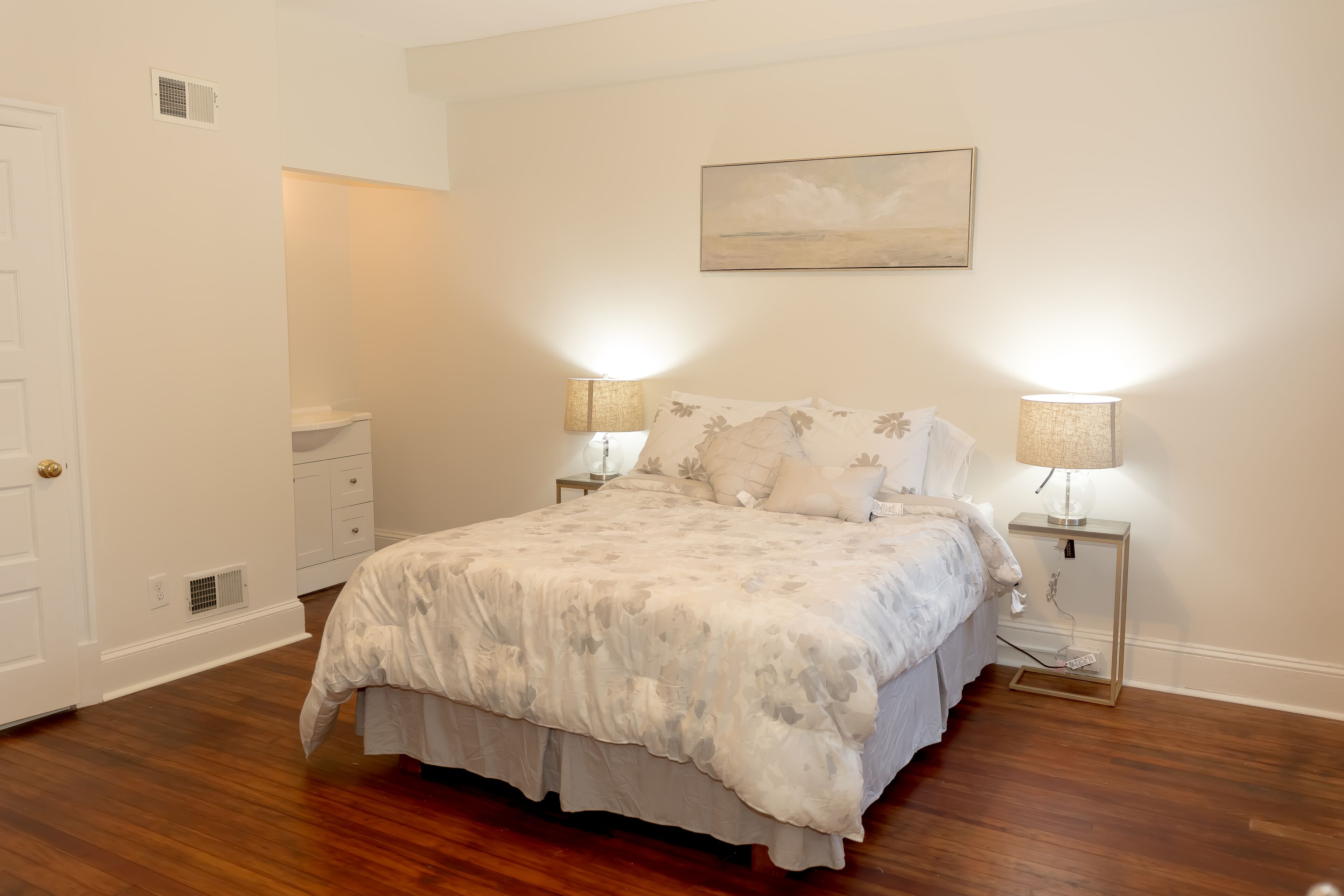 Un dormitorio con una cama grande, dos mesitas de noche con lámparas, una puerta, un armario pequeño y un cuadro en la pared.
