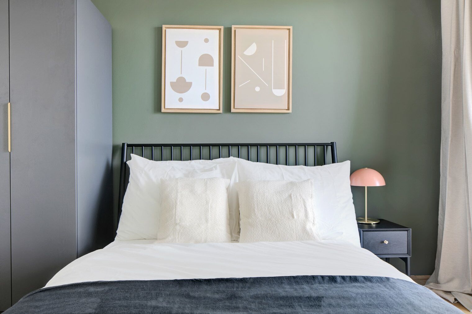 Ein Bett mit weißer Bettwäsche, einem schwarzen Metall-Kopfteil und zwei gerahmten abstrakten Kunstwerken an der Wand.