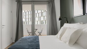 A bedroom with a bed, grey curtains, a window with bars, a small table and chairs, and a wall-mounted lamp. - Modern Wieden 1BR w/ Balcony, Gym, Elevator, walk to U-Bahn, by Blueground (Vienna)