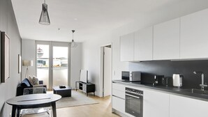 Une cuisine moderne avec des meubles blancs, un plan de travail noir et des appareils électroménagers en acier inoxydable. On y trouve un coin repas avec une table ronde et des chaises, ainsi qu’un canapé à l’arrière-plan.