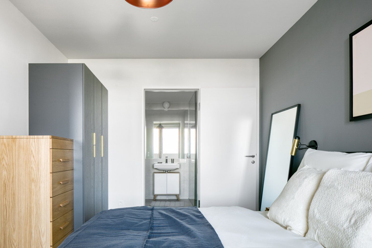 Une chambre à coucher moderne, avec un lit, une commode, un miroir et une salle de bain visible à travers une porte ouverte.